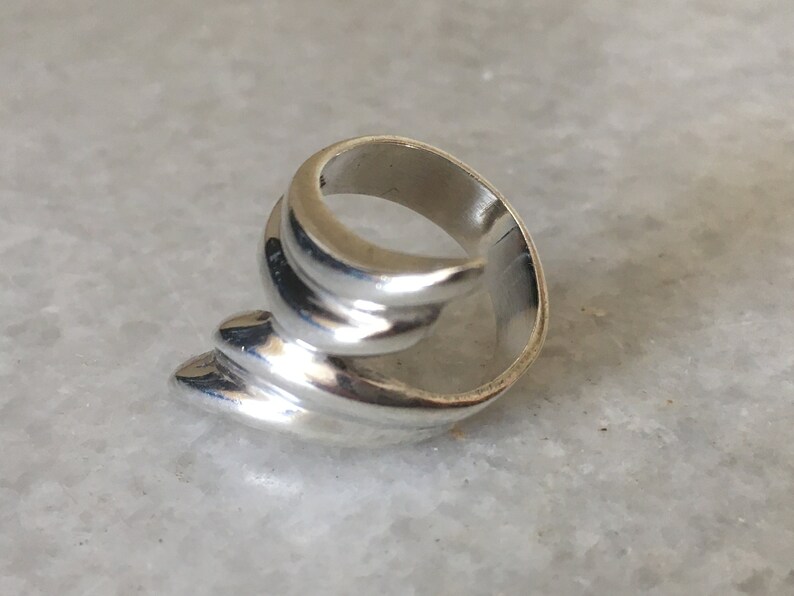 May include: A silver ring with a unique, abstract design. The ring is made of smooth, curved lines that create a flowing, organic shape.