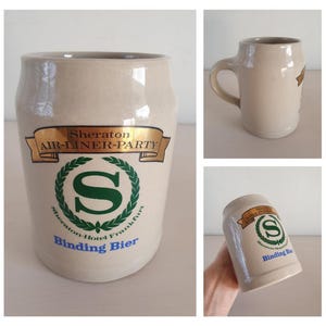 May include: A beige ceramic beer stein with a handle and a cylindrical shape. The stein features a gold banner with the text "Sheraton AIR-DINER-PARTY" and a green emblem with the letter "S". The text "Binding Bier" is also visible.
