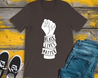 May include: A dark brown t-shirt with a white graphic of a raised fist and the words "BLACK LIVES MATTER" on a banner. The shirt is paired with blue jeans and black sneakers on a yellow and wood-grain surface.