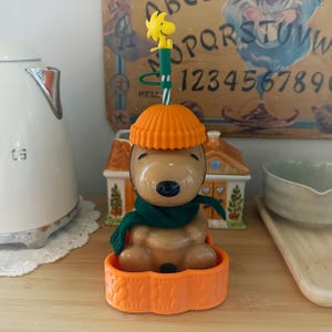May include: A brown Snoopy figure wearing a dark green scarf and an orange ribbed hat, sitting in an orange textured base. A green and white striped straw extends from the top, topped with a small yellow Woodstock figure.