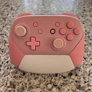 May include: A wireless video game controller in a soft pink hue featuring a white lower panel. The device includes a directional pad, two analog sticks, four action buttons labeled A, B, X, and Y, a home button, plus and minus buttons, and a capture button.
