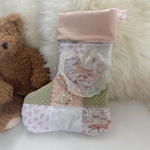 May include: A handmade Christmas stocking with a patchwork design. The stocking features a heart-shaped applique with a deer and floral pattern, accented with lace. The top is a soft pink fabric, and the overall color scheme includes pink, green, and white.