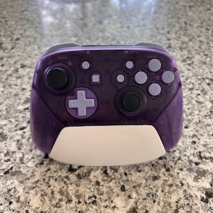 May include: A wireless video game controller featuring a translucent purple plastic shell, two black analog sticks, a light purple directional pad, and light purple action buttons labeled A, B, X, and Y. A solid white panel is attached to the bottom.