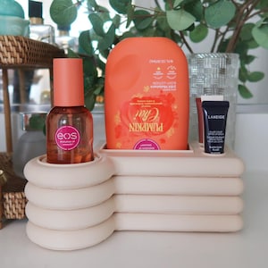 Könnte beinhalten: Ein beigefarbener, gestufter, rechteckiger Organizer mit abgerundeten Kanten enthält eine durchsichtige Flasche mit orangefarbenem Verschluss und eos-Aufschrift, eine große orangefarbene Bodylotion-Flasche Pumpkin Chai und eine kleine schwarze Tube Laneige.