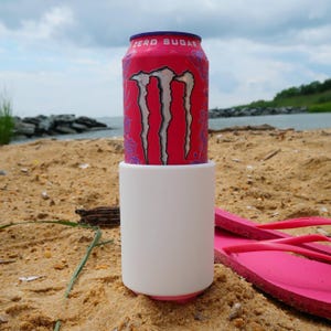 May include: A pink Monster Zero Sugar beverage can inserted into a plain white cylindrical foam can cooler. The can features the iconic black and silver claw logo and the text ZERO SUGAR printed in white near the top rim of the aluminium container.