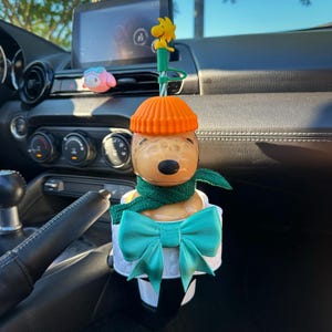 May include: A Snoopy-shaped cup topper featuring a tan dog head wearing an orange ribbed beanie and a dark green knit scarf. A large teal bow is attached to the front of the white cup base. A small yellow Woodstock figure sits atop the green straw.