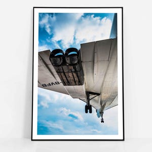 May include: A framed print featuring a low-angle view of an airplane against a blue sky with clouds. The plane's underside and engines are visible, with the registration number -BVF8- on the wing. The image is framed in a black border.