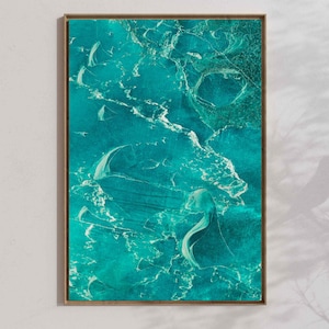May include: Abstract art print featuring a teal and white textured design, framed in a natural wood tone. The artwork has a fluid, organic appearance, resembling water or a geological formation. The piece is mounted on a white wall.