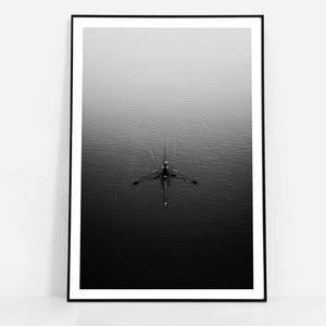 May include: Monochrome framed print featuring a rowing boat on water. The image is captured from above, with the boat centrally positioned and the water reflecting light. The composition is minimalist and tranquil, creating a sense of calm.