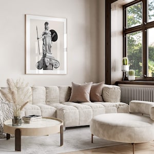 Op de afbeelding: Een witte bank met een zwart-witte ingelijste print van een standbeeld van een vrouw die een speer en schild vasthoudt. De bank staat in een woonkamer met een groot raam en een witte poef.