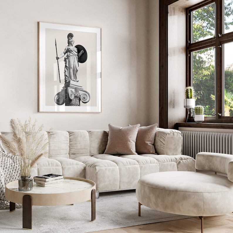 Op de afbeelding: Een witte bank met een zwart-witte ingelijste print van een standbeeld van een vrouw die een speer en schild vasthoudt. De bank staat in een woonkamer met een groot raam en een witte poef.