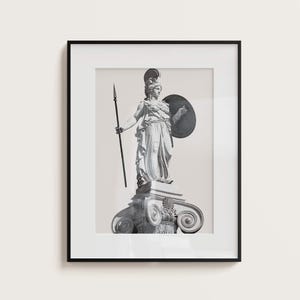 Op de afbeelding: Zwart-witfoto van een standbeeld van een vrouw die een speer en een schild vasthoudt. Het standbeeld staat op een voetstuk met sierlijke details. Het standbeeld is ingelijst in een zwart frame.