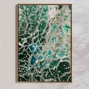 May include: Abstract art print in a wooden frame. The artwork features a blend of green, teal, and silver hues, creating a textured, organic pattern. The design resembles a close-up view of a natural formation.
