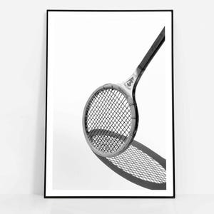Könnte beinhalten: Schwarz-Weiß-Druck in einem Rahmen mit einem Vintage-Tennisschläger. Der Schläger ist abgewinkelt und wirft einen Schatten auf den weißen Hintergrund. Der Rahmen ist schwarz, und der Schläger hat einen dunklen Griff und einen hellen Kopf mit Netz.