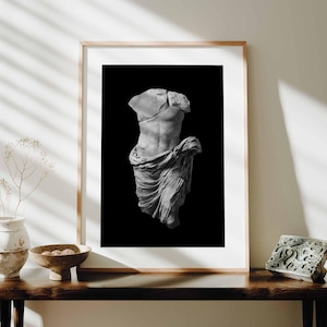 May include: Black and white print of a classical sculpture of a torso, framed in a light wood frame. The sculpture is set against a black background. The artwork is displayed on a dark wood table.