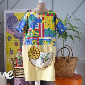 以下が含まれることがあります： 黄色と青のパッチワークドレスに、黄色と白のクロシェのポケットが付いている。ドレスは白いマネキンに白いスタンドで置かれている。ドレスは半袖で、丸いネックライン。ドレスは木製の床に敷かれたラグの上にある。