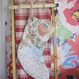 Könnte beinhalten: Ein gesteppter Weihnachtsstrumpf mit einem Patchwork-Design aus Blumen- und Pflanzenmustern in den Farben Rosa, Grün, Blau und Creme. Der Strumpf hängt an einem Haken an einer Holzleiter, die mit einem rot-weiß karierten Stoff behangen ist.