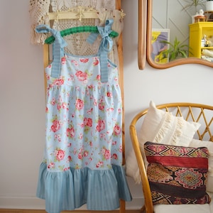 Könnte beinhalten: Ein hellblaues Blumenkleid mit einem Rüschen-Saum und blau-weiß gestreiften Trägern, das an einem Holzgestell hängt.