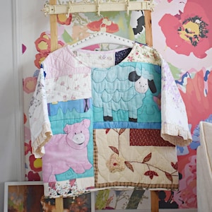 May include: A patchwork quilt top with a pink pig, a blue sheep, and floral patterns. The quilt is hanging on a wooden ladder against a floral wallpaper.