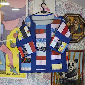 May include: A blue patchwork shirt with a variety of colorful fabric squares. The shirt has a round neckline and three-quarter sleeves. The shirt is hanging on a hanger against a floral wallpaper background.