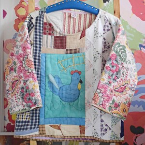 Könnte beinhalten: Ein Patchwork-Deckenoberteil mit einem blauen Hühnermotiv. Die Decke besteht aus verschiedenen Stoffen in den Farben Rosa, Blau, Weiß und Braun. Die Decke hängt an einem Holzaufhänger.