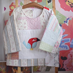 Pode incluir: Uma camisa branca acolchoada com um design de patchwork. A camisa tem mangas compridas e um decote redondo. As mangas são feitas de diferentes tecidos estampados, incluindo listras, xadrez e estampas florais. Uma aplicação de pássaro azul e vermelho está centrada na frente da camisa.