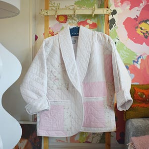 Könnte beinhalten: Ein gesteppter weißer Bademantel mit rosa Akzenten hängt an einer Holzleiter. Der Bademantel hat ein Rautenmuster, einen Schalkragen und rosa quadratische Taschen. Der Hintergrund umfasst eine Blumentapete und eine dekorative Leiter.