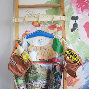 May include: A colorful, patchwork top with map and landscape prints, hanging on a blue hanger. The sleeves are brown with text. The top is displayed on a wooden ladder against a floral wallpaper background.