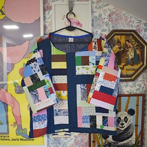 Puede incluir: Una camisa azul de patchwork hecha con una variedad de telas coloridas. La camisa tiene un cuello redondo y mangas tres cuartos. La camisa est&aacute; colgada de una percha contra un fondo de papel tapiz floral.