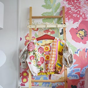 Op de afbeelding: Een wit, bloemig geprint crop top met een gele elastische tailleband hangt aan een houten ladder tegen een achtergrond van bloemenbehang. Het topje is gemaakt van verschillende stofresten die aan elkaar zijn genaaid, waardoor een patchwork-ontwerp ontstaat.