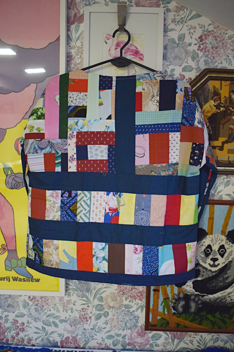 Puede incluir: Una camisa azul de patchwork con una variedad de telas y patrones coloridos. La camisa est&aacute; colgada de una percha y tiene una forma cuadrada.