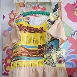 Peut inclure: Une chemise vintage jaune et marron avec un imprimé de calendrier et une scène de plage avec un bateau. La chemise a un col à volants et des manches courtes avec un tissu superposé de couleur pêche. La chemise est suspendue à un cintre blanc.