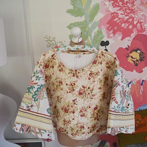 May include: A cream-colored, floral-print top with elbow-length sleeves. The top features a round neckline and is displayed on a white mannequin. The sleeves have a contrasting patterned fabric with green, blue, and red accents. The background includes floral wallpaper.