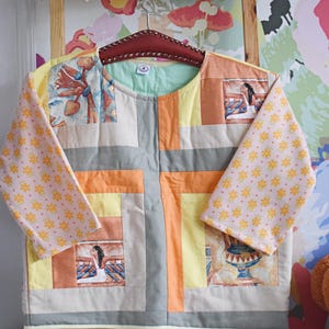 May include: A patchwork jacket with a round neckline and long sleeves. The jacket features a mix of colors including yellow, orange, gray, and beige. The sleeves have a pink floral pattern. The jacket is hanging on a wooden hanger.