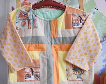 Puede incluir: Chaqueta patchwork con cuello redondo y mangas largas. La chaqueta presenta una mezcla de colores como amarillo, naranja, gris y beige. Las mangas tienen un estampado floral rosa. La chaqueta está colgada en una percha de madera.