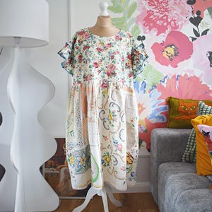 以下が含まれることがあります： マネキンに着せられた花柄のドレス。 ドレスは、小さな花柄のクリーム色のボディスと、青いトリムのフリル袖が特徴です。 スカートは、花柄と模様の生地のパッチワークです。 ドレスは白いスタンドに展示されています。