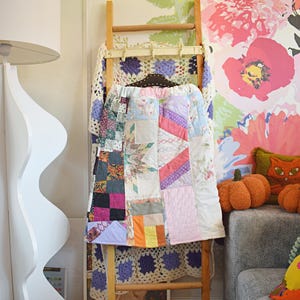 May include: A wooden ladder displays a patchwork quilt and a crocheted blanket. The quilt features various fabric patterns and colours, including pink, purple, and orange. The background includes floral wallpaper and decorative pumpkins.