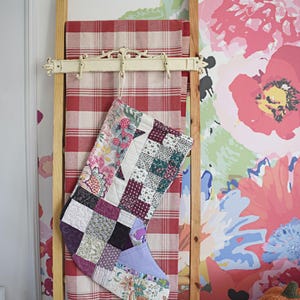 Puede incluir: Un calcetín navideño acolchado con un diseño de patchwork de varios patrones de tela florales y geométricos en tonos morado, rosa, verde y blanco, colgado de un gancho de metal blanco en una escalera de madera cubierta con una tela de cuadros rojos y blancos.