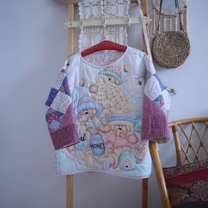 Pode incluir: Uma jaqueta acolchoada branca com um design de patchwork com ursos e potes de mel. A jaqueta está pendurada em uma escada de madeira com um chapéu marrom, uma renda branca de crochê e uma cesta de vime.