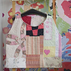 Pode incluir: Um colete patchwork com uma variedade de padrões e cores de tecido, incluindo rosa, vermelho e amarelo. O colete está pendurado em uma escada de madeira contra um fundo de papel de parede floral. O colete apresenta um patch em forma de coração.