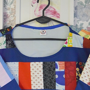 Puede incluir: Una camisa azul de patchwork con una variedad de patrones coloridos, incluyendo dise&ntilde;os florales, geom&eacute;tricos y abstractos. La camisa tiene un escote redondo y est&aacute; colgada de una percha negra.