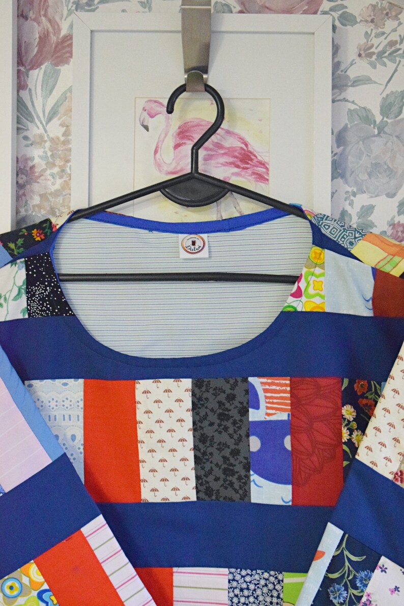 Puede incluir: Una camisa azul de patchwork con una variedad de patrones coloridos, incluyendo dise&ntilde;os florales, geom&eacute;tricos y abstractos. La camisa tiene un escote redondo y est&aacute; colgada de una percha negra.