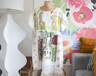 Puede incluir: Vestido blanco de manga corta con un estampado único. La parte superior presenta figuras en verde, amarillo y naranja, con el año "1998" impreso debajo. La parte inferior muestra un estampado de un pueblo pintoresco. El vestido está en un maniquí.