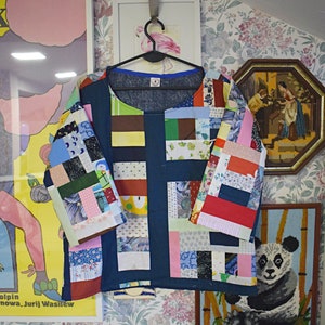 Puede incluir: Una camisa azul de patchwork con una variedad de cuadrados de tela de colores. La camisa tiene un cuello redondo y mangas cortas. La camisa est&aacute; colgada de una percha contra un fondo de papel tapiz floral.