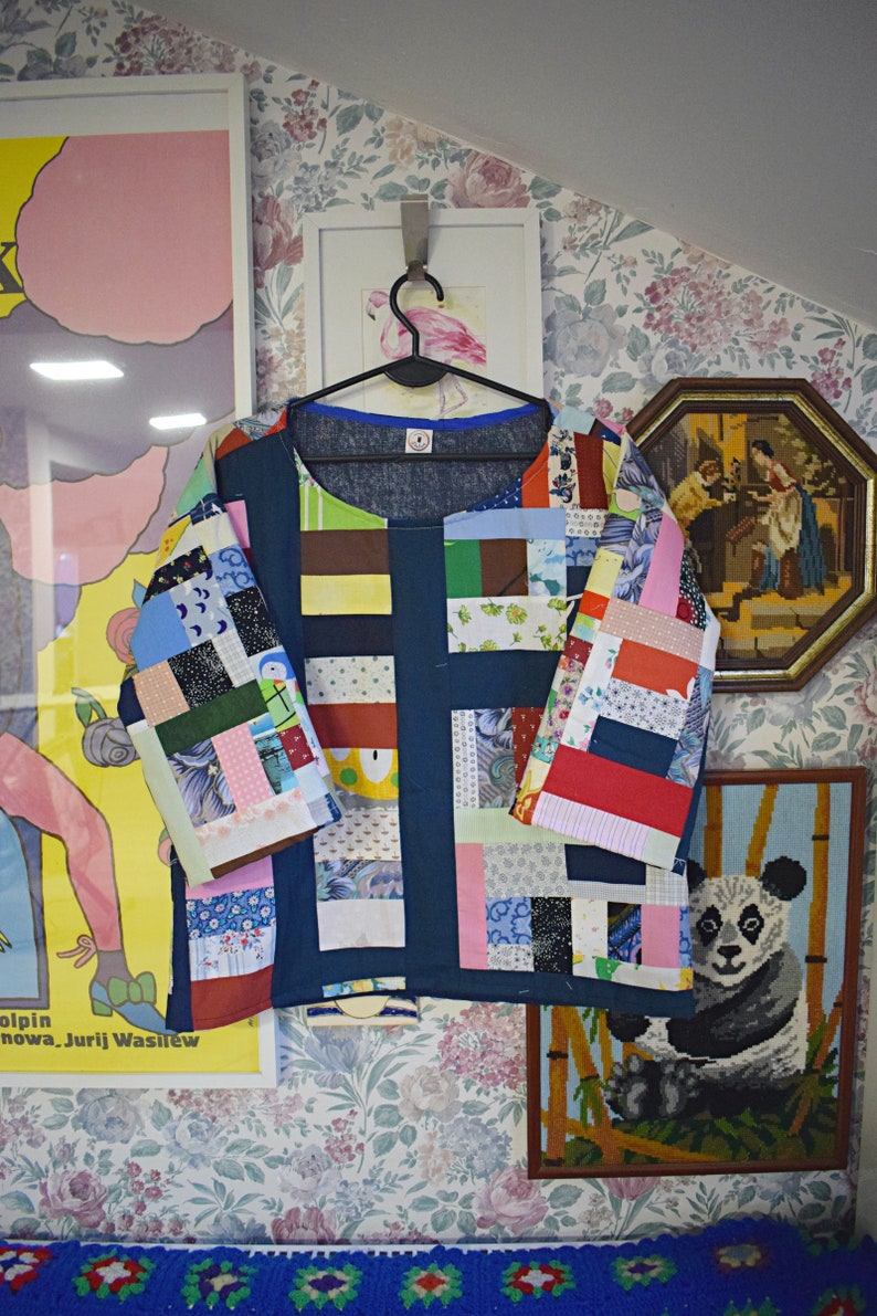 Puede incluir: Una camisa azul de patchwork con una variedad de cuadrados de tela de colores. La camisa tiene un cuello redondo y mangas cortas. La camisa est&aacute; colgada de una percha contra un fondo de papel tapiz floral.