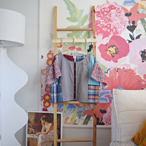 Pode incluir: Uma camisa de patchwork com vários padrões de xadrez em azul, vermelho e branco pendurada em uma escada de madeira contra um fundo de papel de parede floral.