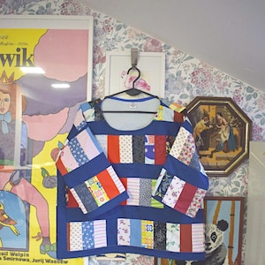 Puede incluir: Una camisa de manga larga de patchwork azul y blanco colgada de una percha. La camisa est&aacute; hecha de diferentes cuadrados de tela de colores cosidos juntos. La camisa est&aacute; colgada frente a una pared con papel tapiz floral.