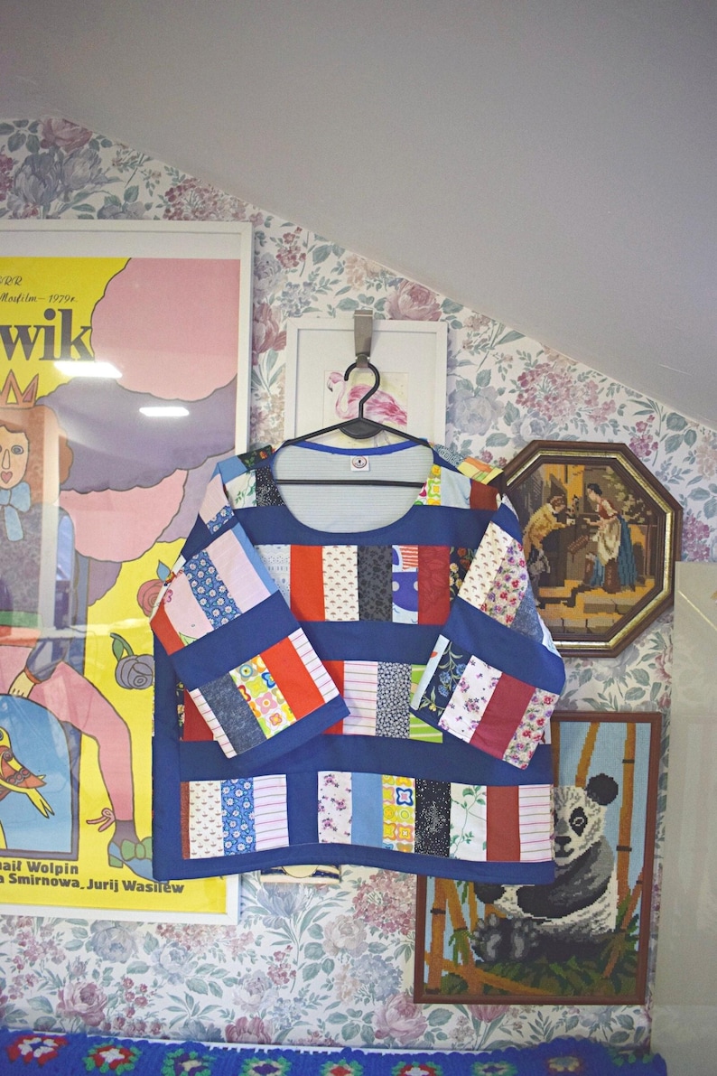 Puede incluir: Una camisa de manga larga de patchwork azul y blanco colgada de una percha. La camisa est&aacute; hecha de diferentes cuadrados de tela de colores cosidos juntos. La camisa est&aacute; colgada frente a una pared con papel tapiz floral.