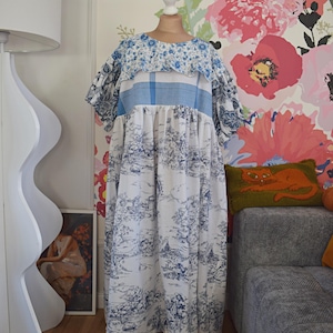 Op de afbeelding: Een lange jurk met een blauw-witte toile de Jouy-print, met een gerimpelde halslijn en korte mouwen. De jurk wordt gepresenteerd op een mannequin. De achtergrond heeft bloemenbehang. De lengte van de jurk is zichtbaar.