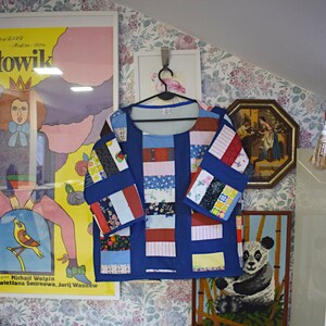 May include: A blue patchwork shirt with long sleeves and a crew neck. The shirt is made of different fabrics and patterns, including stripes, florals, and polka dots. The shirt is hanging on a hanger against a floral wallpaper background.